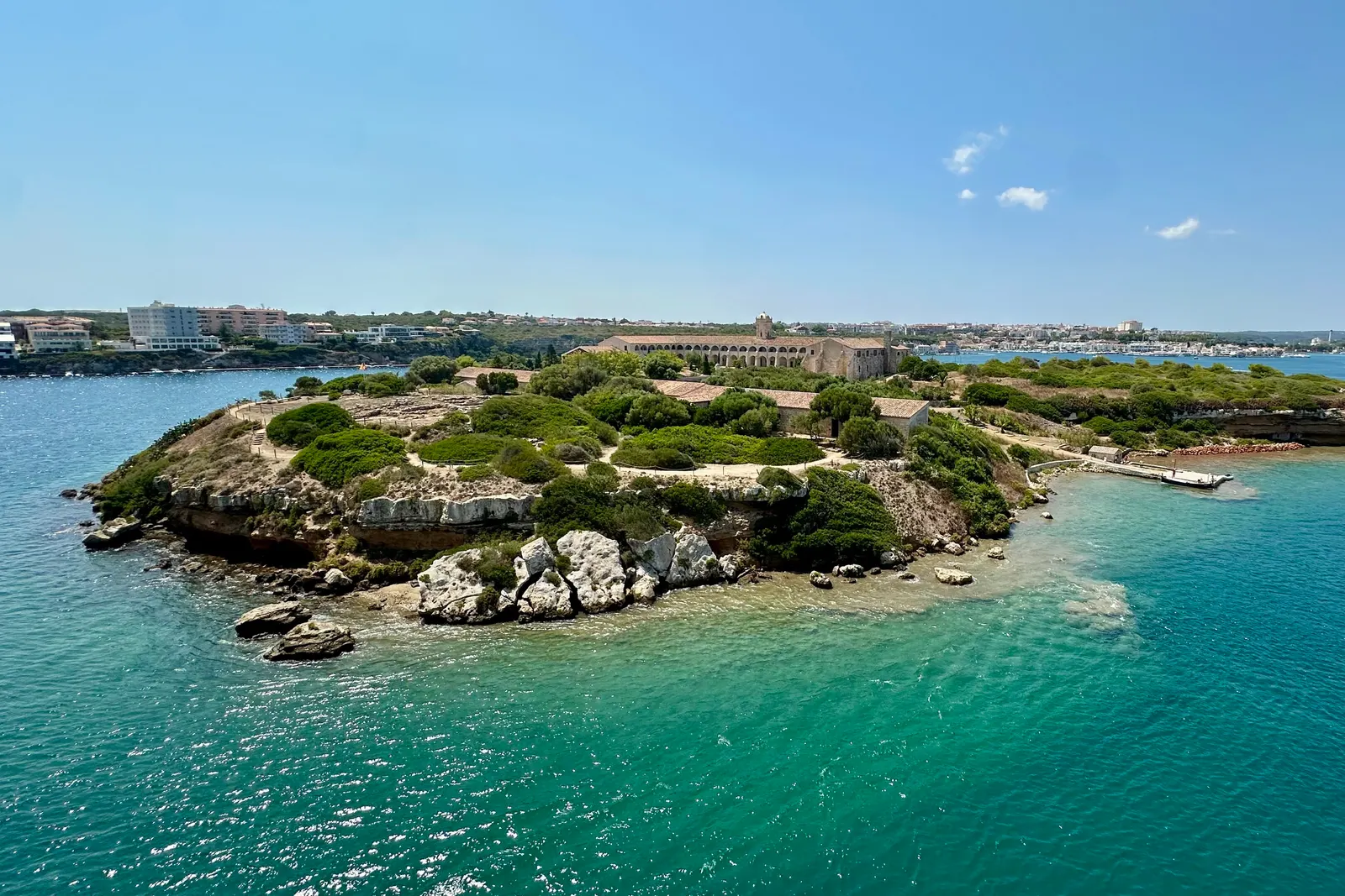 La Illa del Rei en el puerto de Maó, rodeada de agua turquesa, con la costa de la bahía al fondo