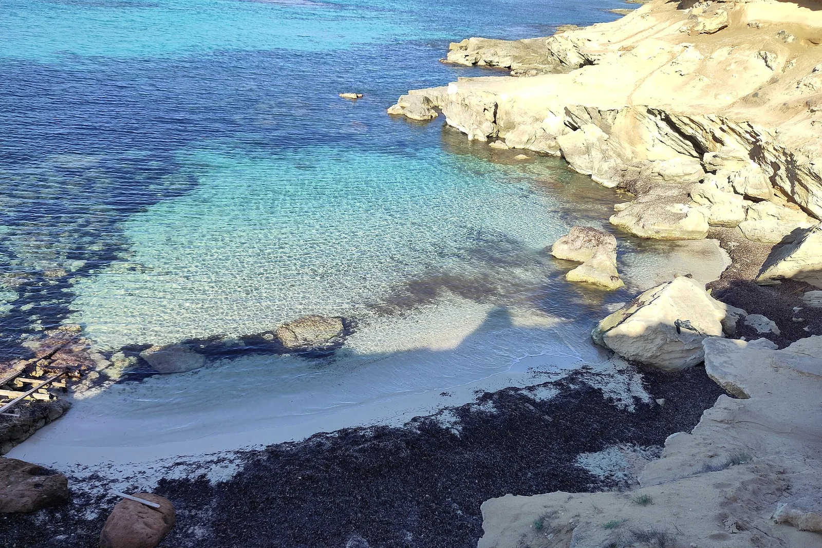 El agua transparente del Caló des Mort sobre la arena clara, entre plataformas de roca dorada y sin nadie en la orilla.