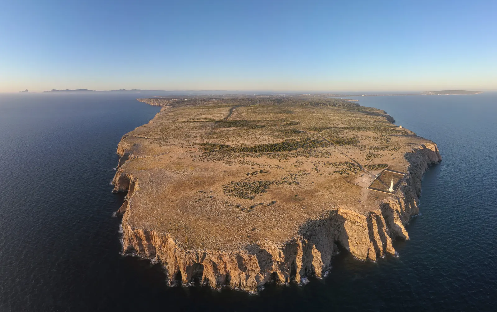 Vista aérea del Cap de Barbaria: la meseta árida, los acantilados sobre el mar y el faro blanco en el extremo