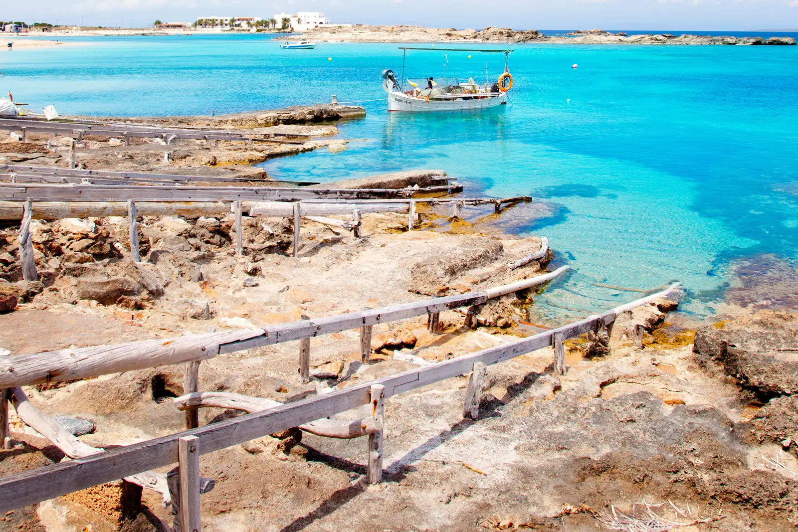 Varaderos de madera sobre la roca y un llaüt fondeado en agua turquesa, con casas blancas al fondo, en la costa de Formentera.