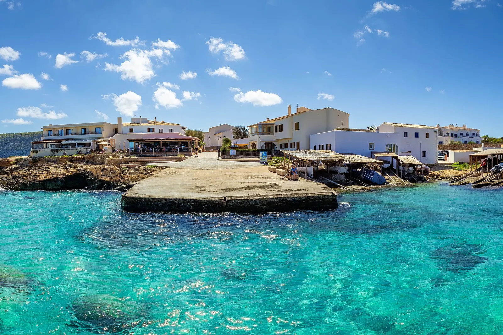 El pequeño puerto de es Caló de Sant Agustí, en Formentera: agua turquesa transparente, un muelle de piedra, casas blancas con la terraza de un restaurante junto al mar y casetas de madera para las barcas.
