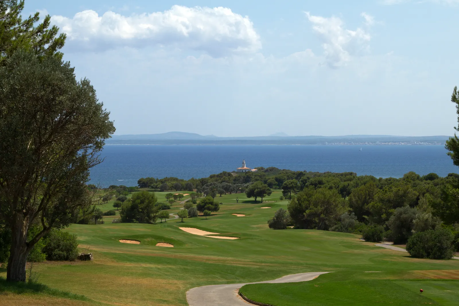 Calle del Club de Golf Alcanada entre pinos y búnkeres, con la bahía de Alcúdia y el faro del islote de Alcanada al fondo.