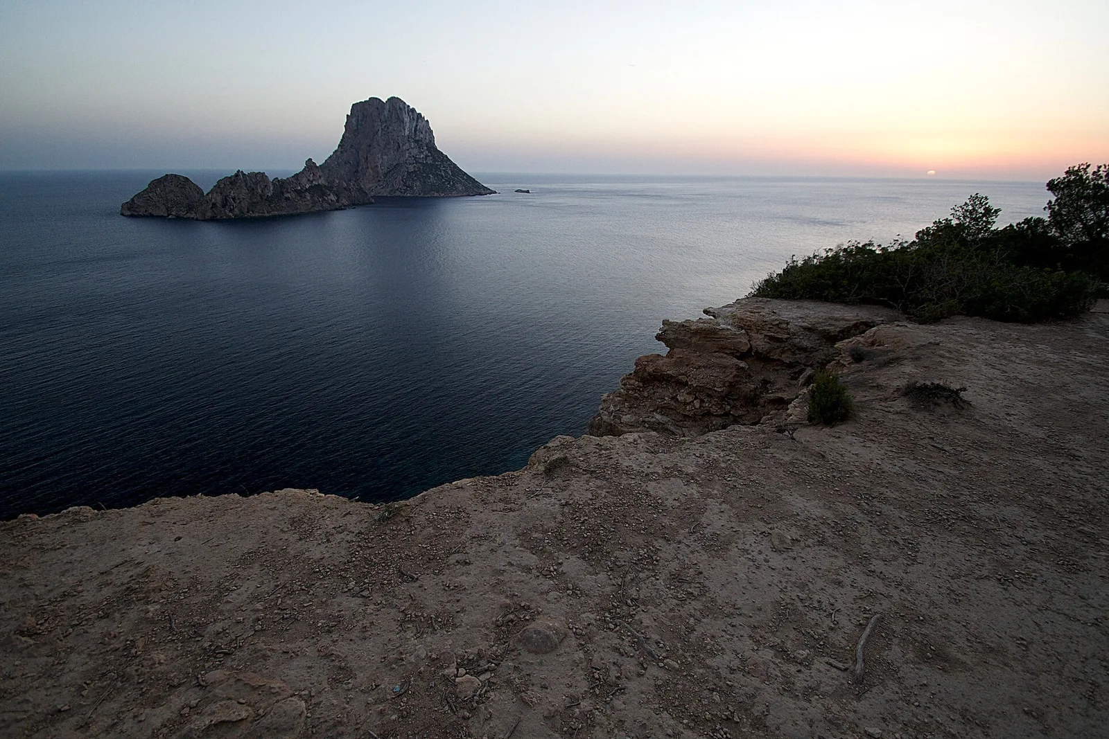 Es Vedrà recortado sobre un mar liso al atardecer, visto desde el borde rocoso de la costa de Cala d'Hort.