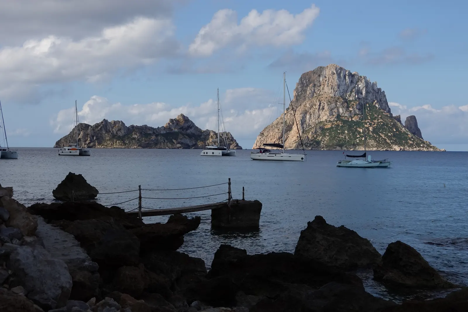 Es Vedrà y es Vedranell desde la costa, con veleros fondeados y rocas en primer plano, al final de la tarde