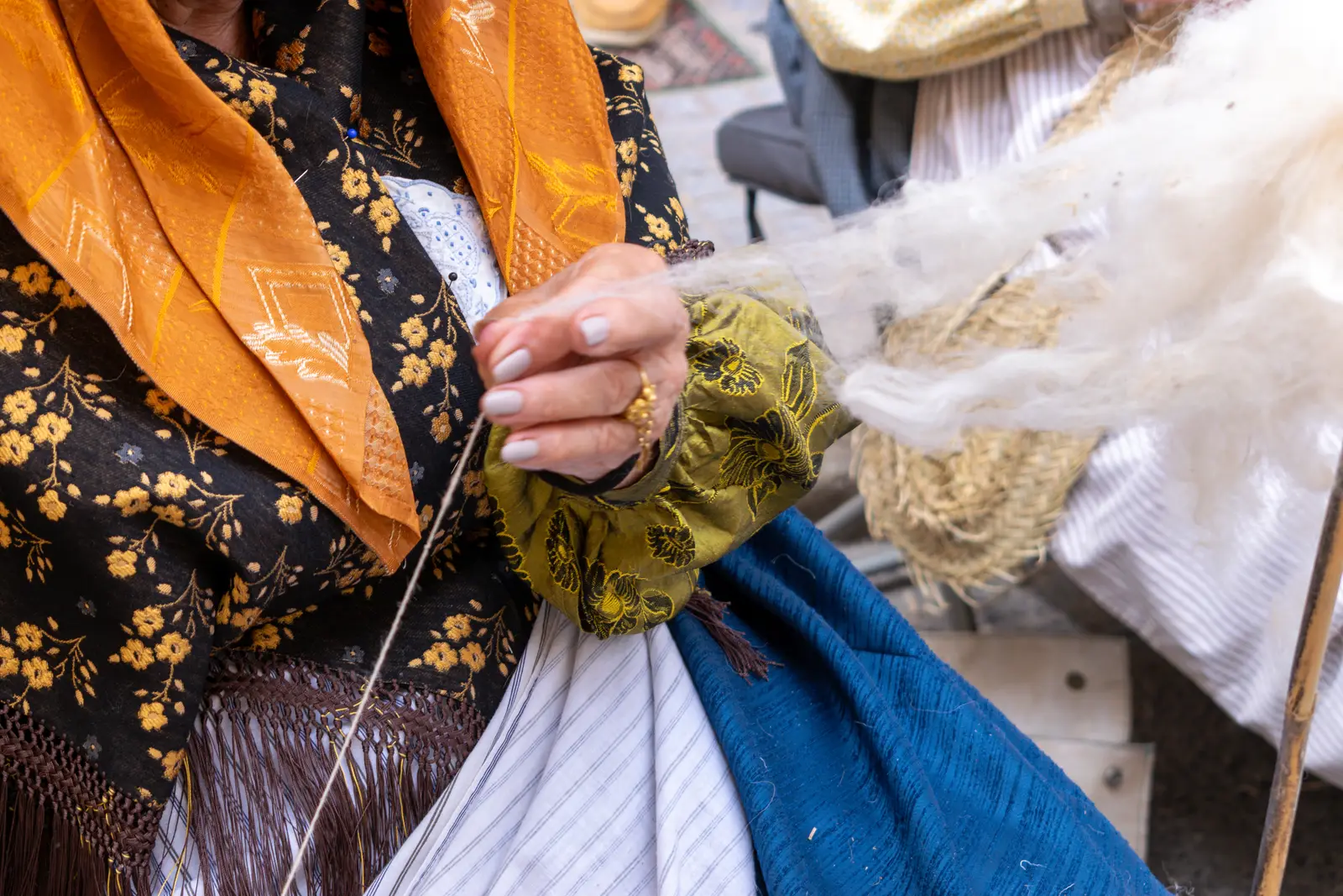 Manos de una mujer con traje tradicional ibicenco hilando lana en un mercado, con el mantón bordado y el delantal a la vista.