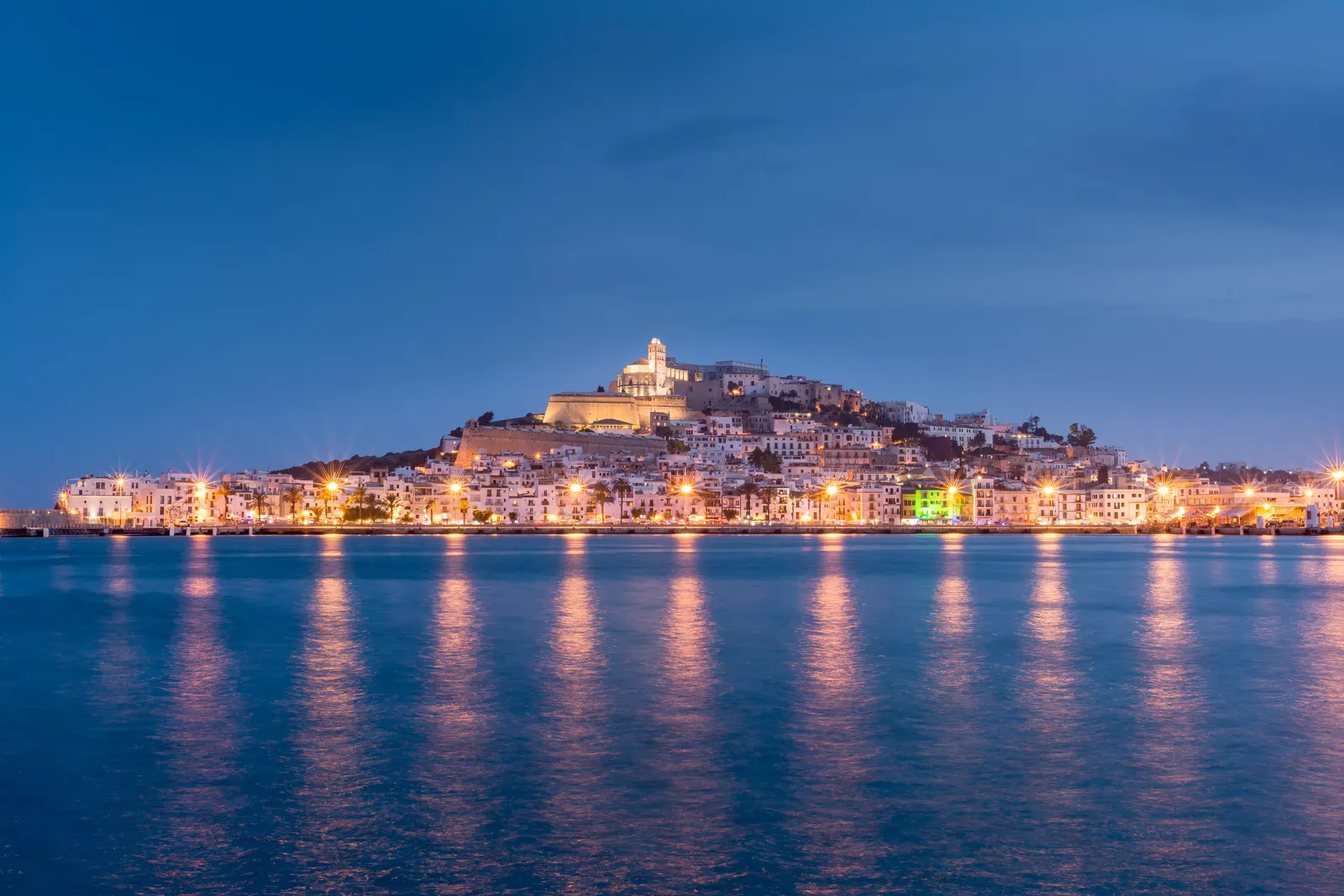 Dalt Vila iluminada al anochecer vista desde el mar, con las luces del paseo reflejadas en el agua en calma.