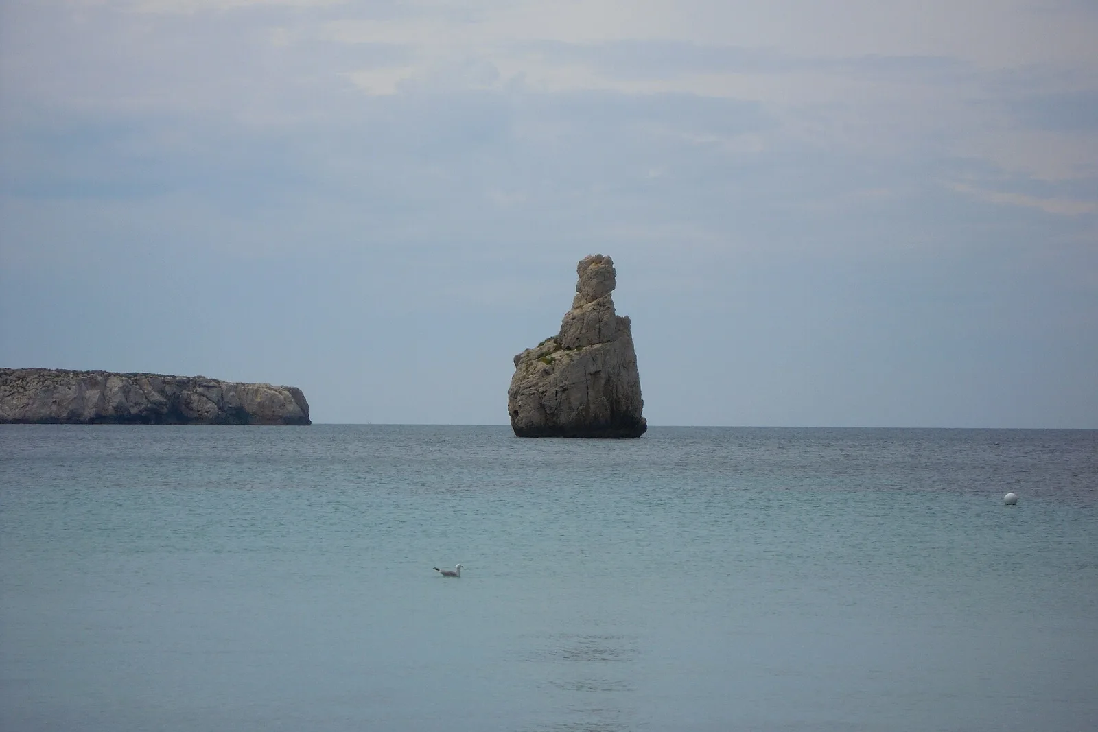 El islote de Cap Bernat frente a la cala de Benirràs, con el mar en calma, el cielo blanco y una gaviota posada en el agua.