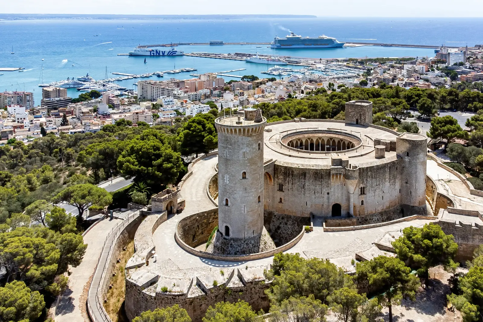 Vista aérea del castillo de Bellver sobre su colina de pinos, con la bahía y el puerto de Palma al fondo.