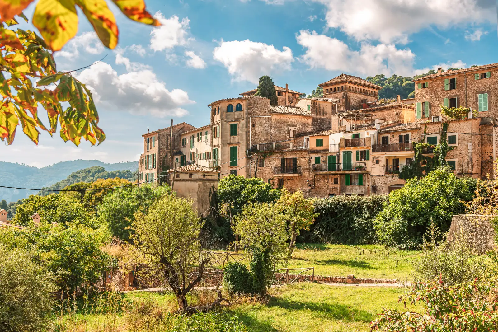 Valldemossa entre árboles, con las casas de piedra dorada en la ladera y hojas otoñales en primer plano.