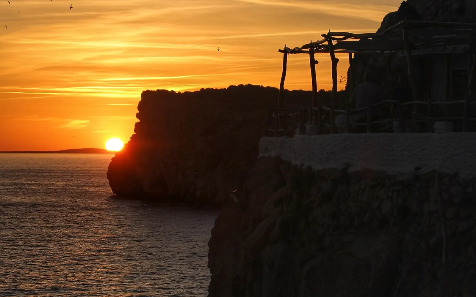 las terrazas de la Cova d'en Xoroi recortadas sobre un atardecer dorado en el acantilado de Cala'n Porter, Menorca