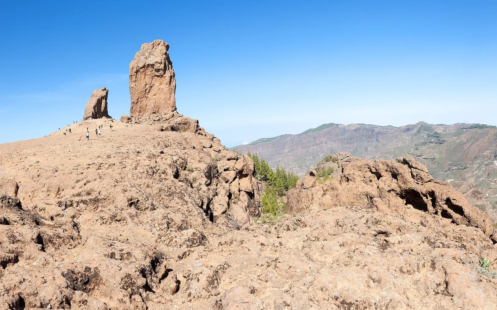 El Roque Nublo se recorta sobre la cumbre rocosa, frente a la gran caldera de Tejeda y sus montañas bajo un cielo despejado.