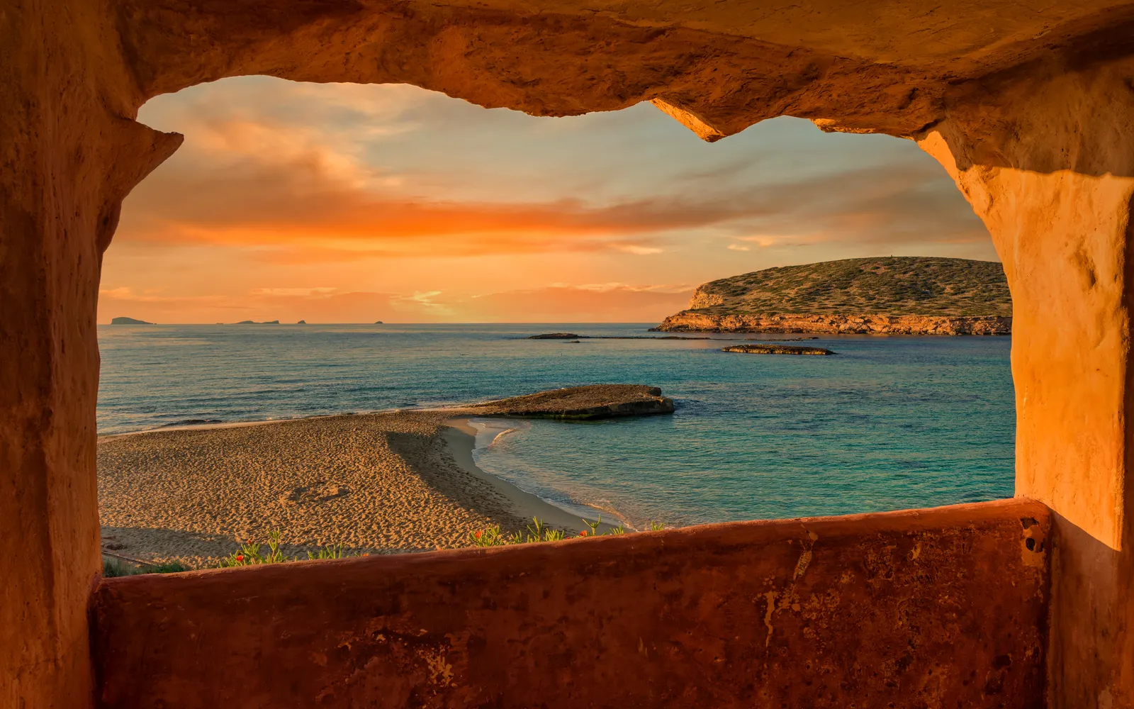 Atardecer anaranjado sobre una cala de la costa de poniente de Ibiza, visto desde el hueco de un muro de piedra.