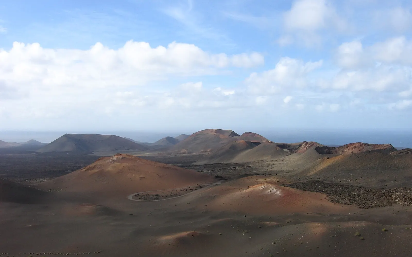 Conos volcánicos rojizos y campos de ceniza de Timanfaya se extienden hasta el mar bajo un cielo de nubes blancas.
