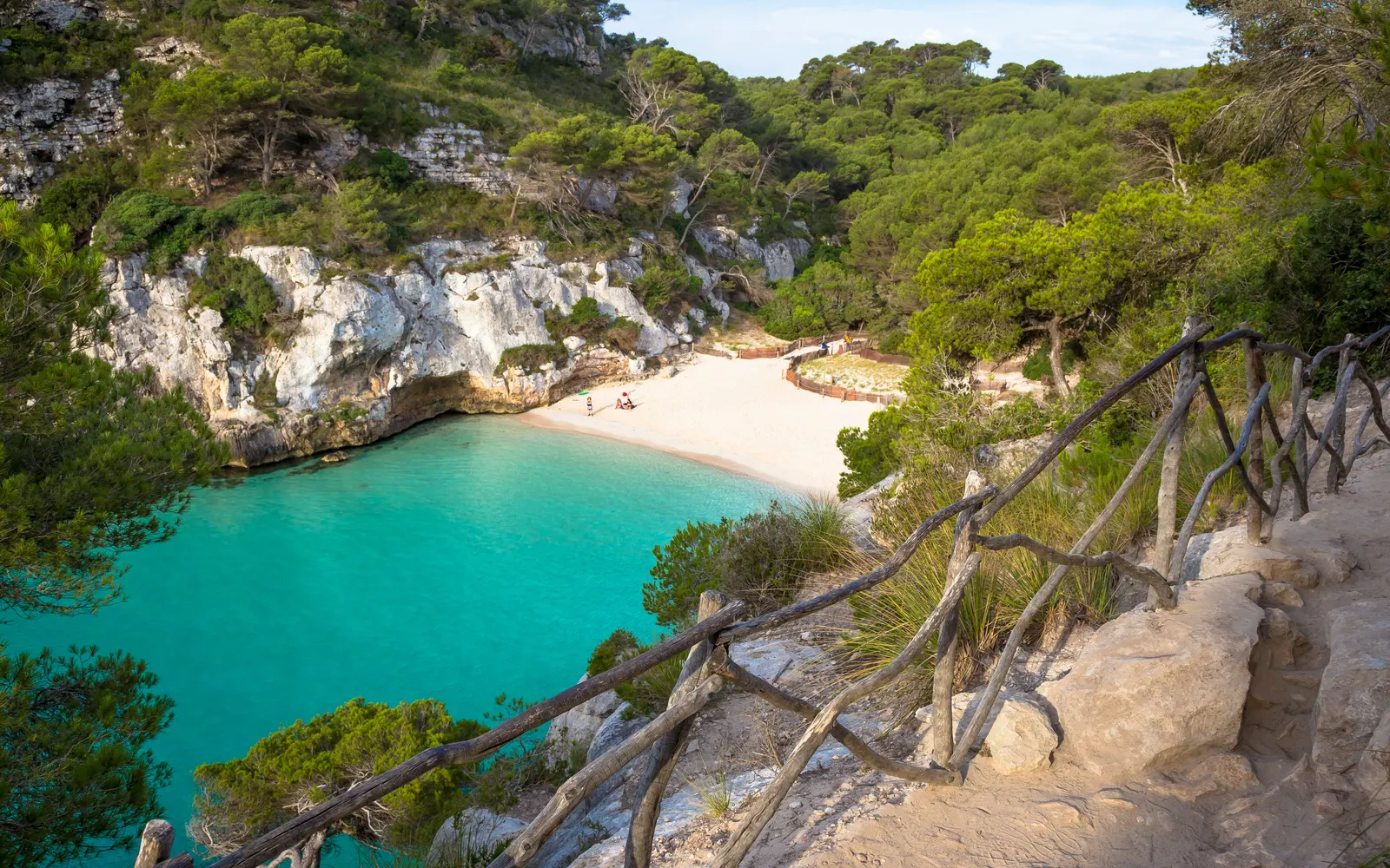 Cala de aguas turquesa entre pinos, con una barandilla de madera en el camino de bajada, en la costa sur de Menorca.