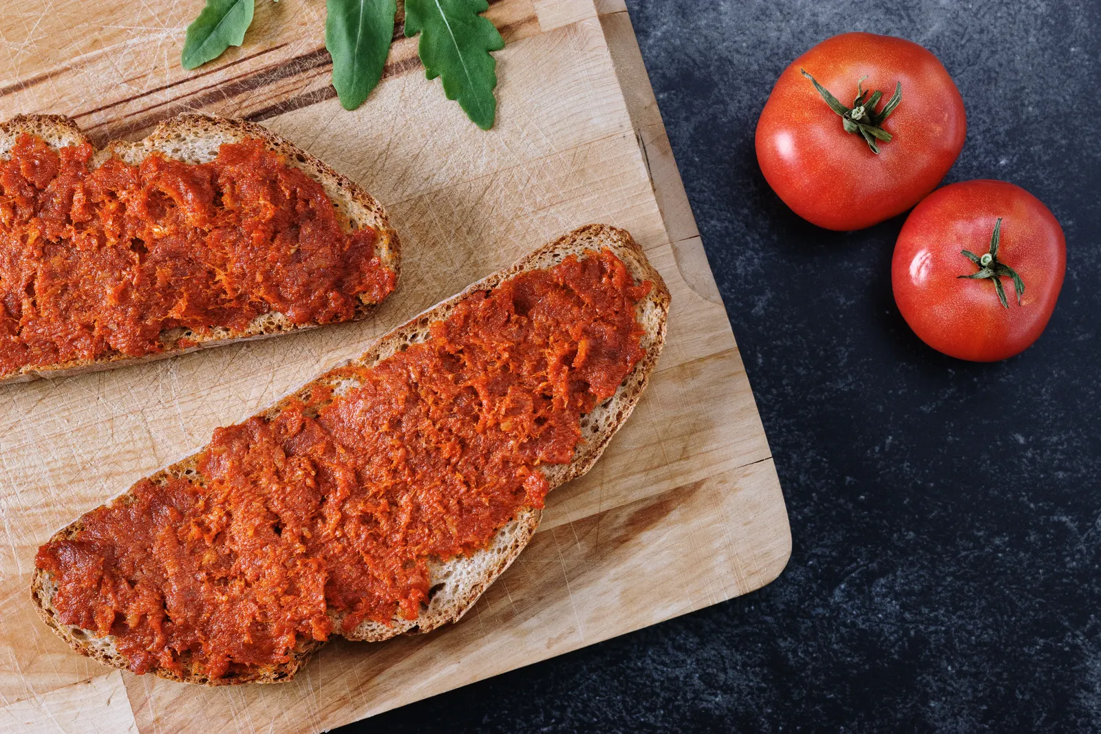 Rebanadas de pan rústico untadas con sobrasada sobre una tabla de madera, junto a unos tomates.