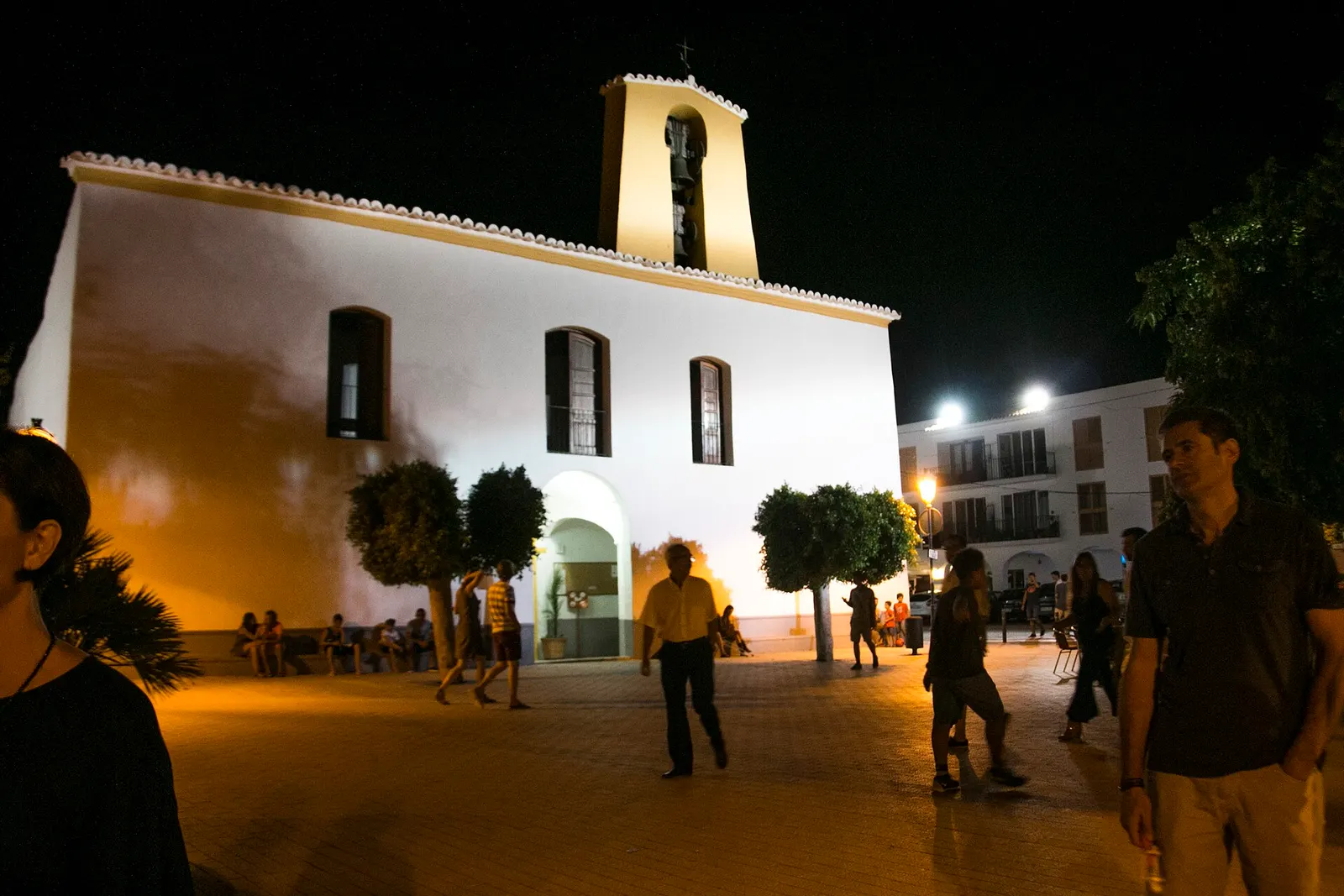 La iglesia blanca de Santa Gertrudis iluminada una noche de verano, con gente en la plaza