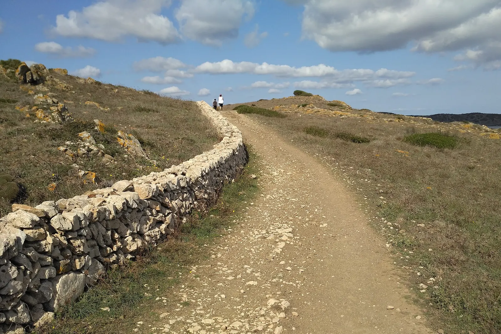 Il Camí de Cavalls verso Binillautí, uno sterrato accanto a un muretto a secco, con un escursionista in lontananza