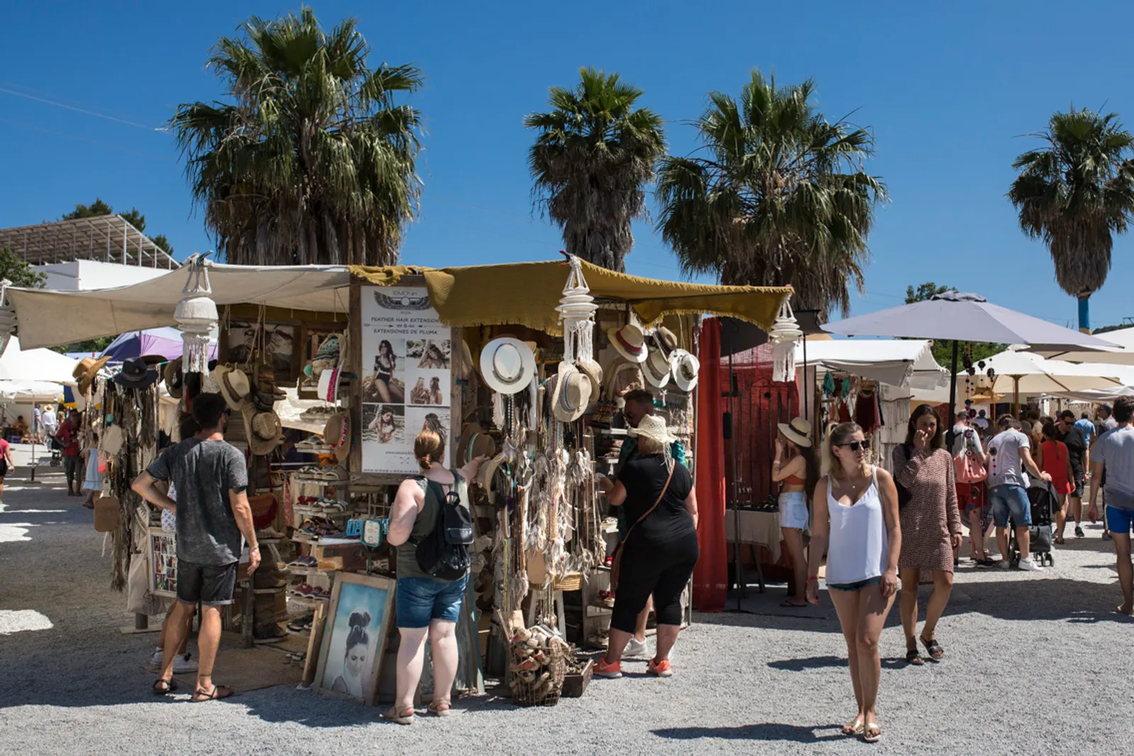 Puestos del mercadillo de Las Dalias con sombreros y artesanía, bajo las palmeras, con visitantes