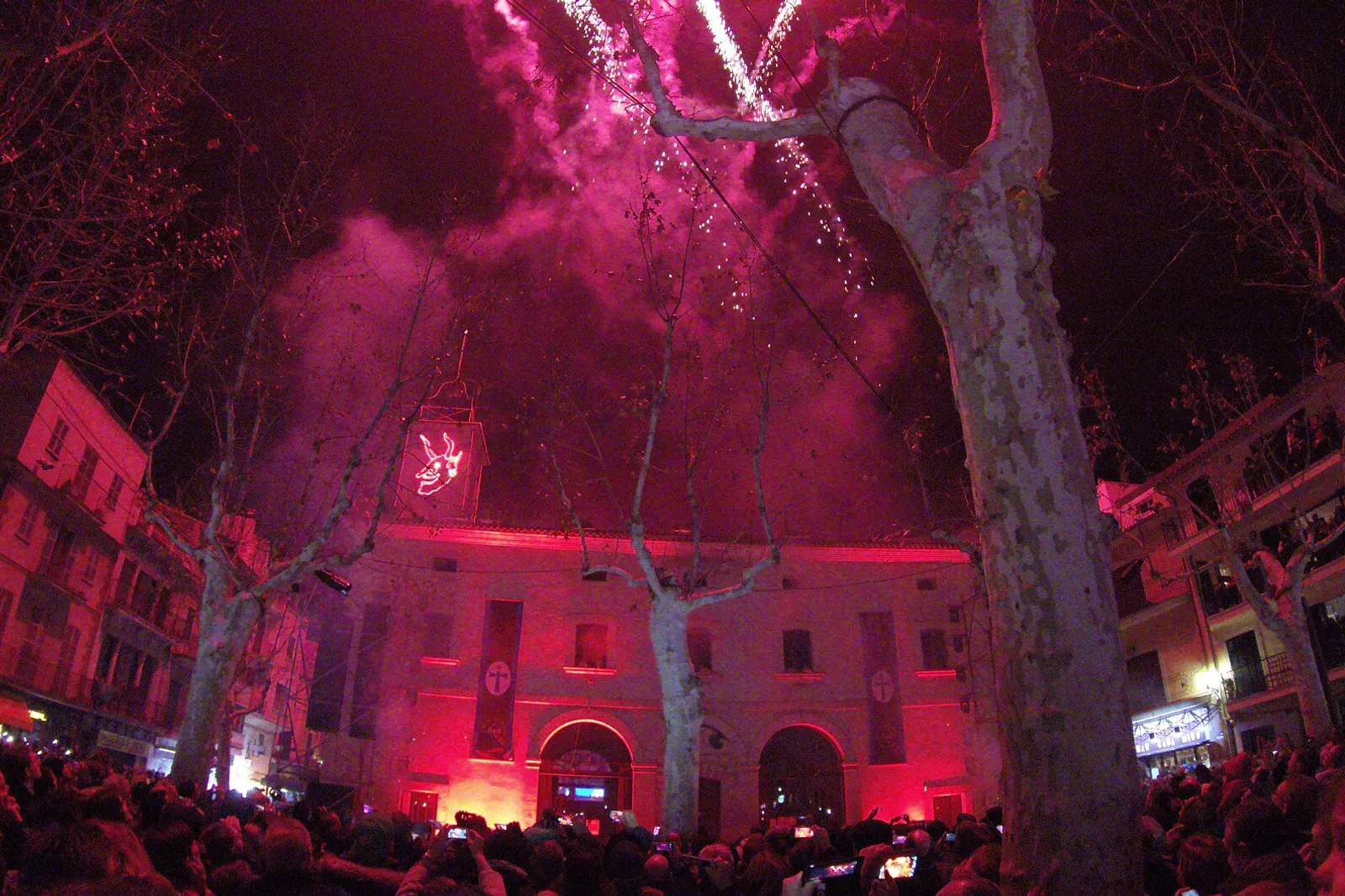 Fuochi d'artificio rossi e rosa sopra la facciata illuminata durante lo spettacolo piromusicale di Sant Antoni de Sa Pobla, con il pubblico davanti