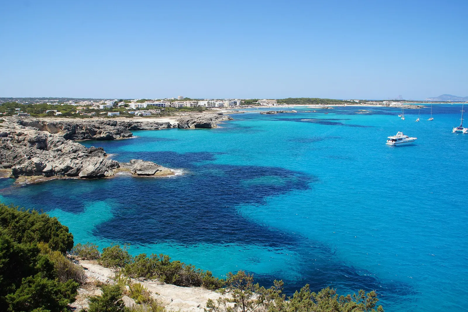 La costa de Es Pujols con el agua turquesa entre las rocas y el pueblo al fondo