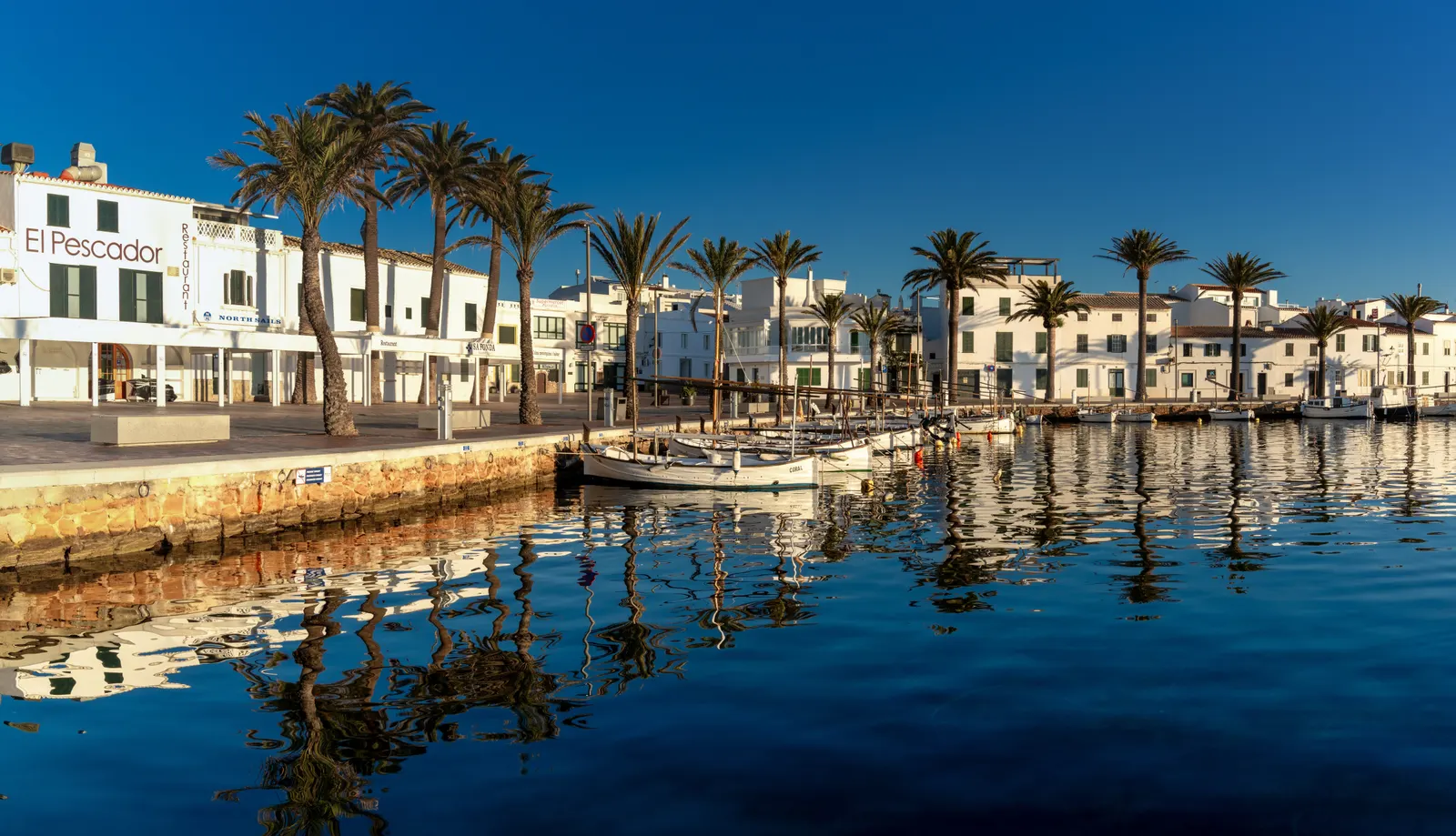 El paseo de Fornells reflejado en el agua quieta de la bahía: casas blancas, palmeras y un llaüt amarrado.