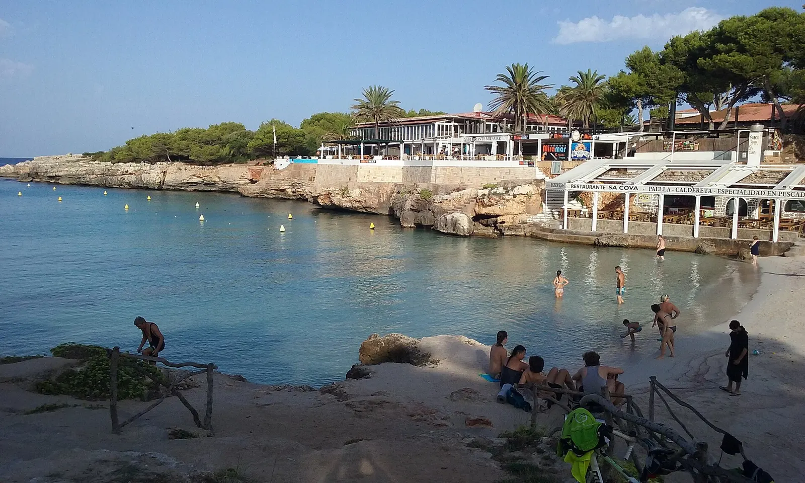 Cala Blanca con bañistas en la orilla y las terrazas asomadas sobre la roca