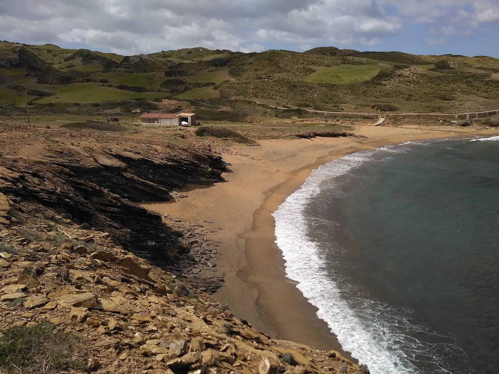 Cala Mica desde lo alto: una lengua de arena y el oleaje rompiendo en la orilla