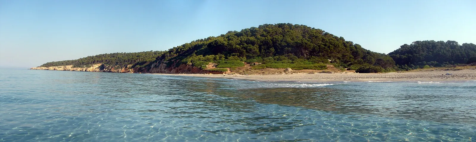 La playa de Binigaus con el promontorio verde y el agua clara batiendo la arena