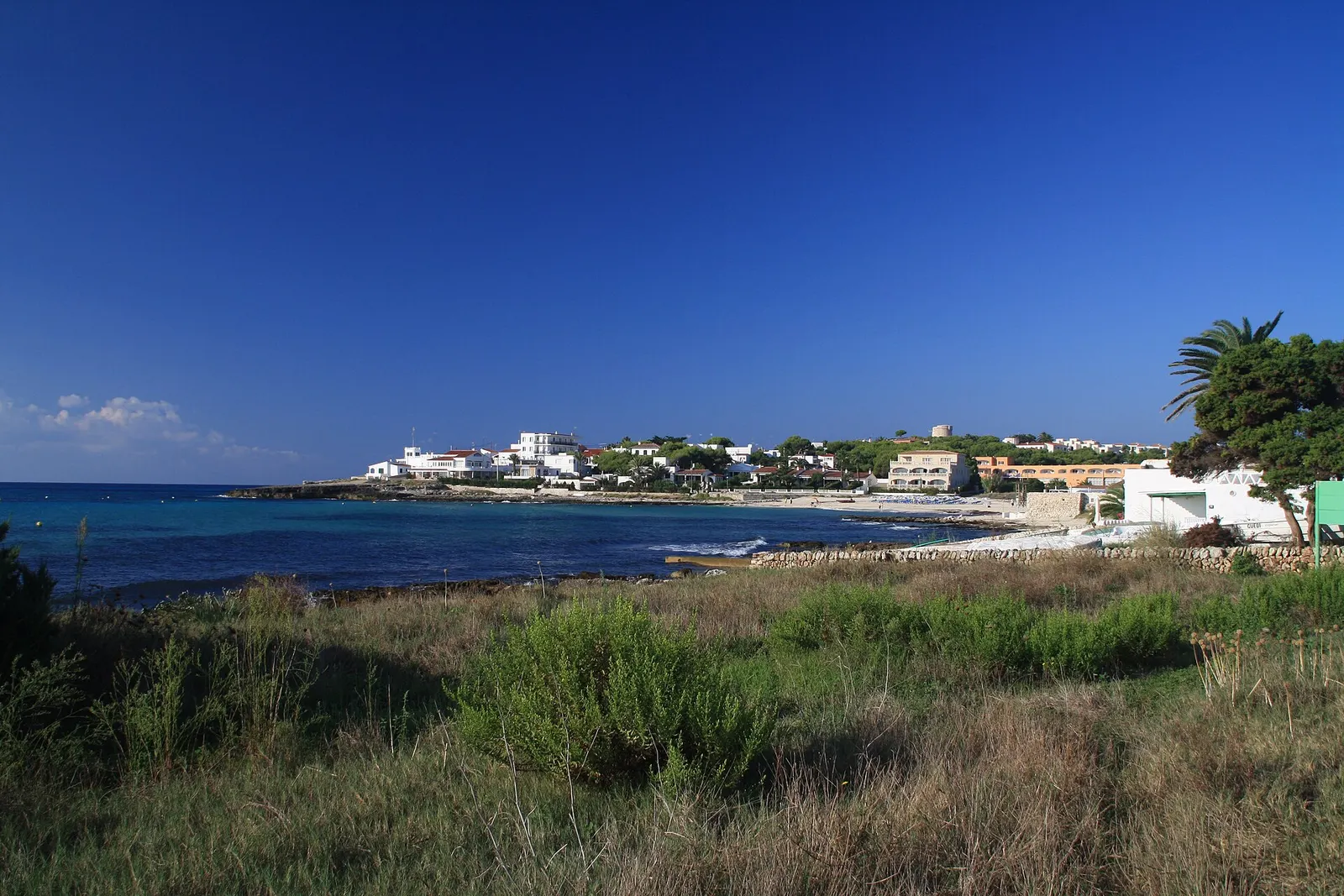 O casario branco de Punta Prima junto ao mar, visto entre o mato baixo