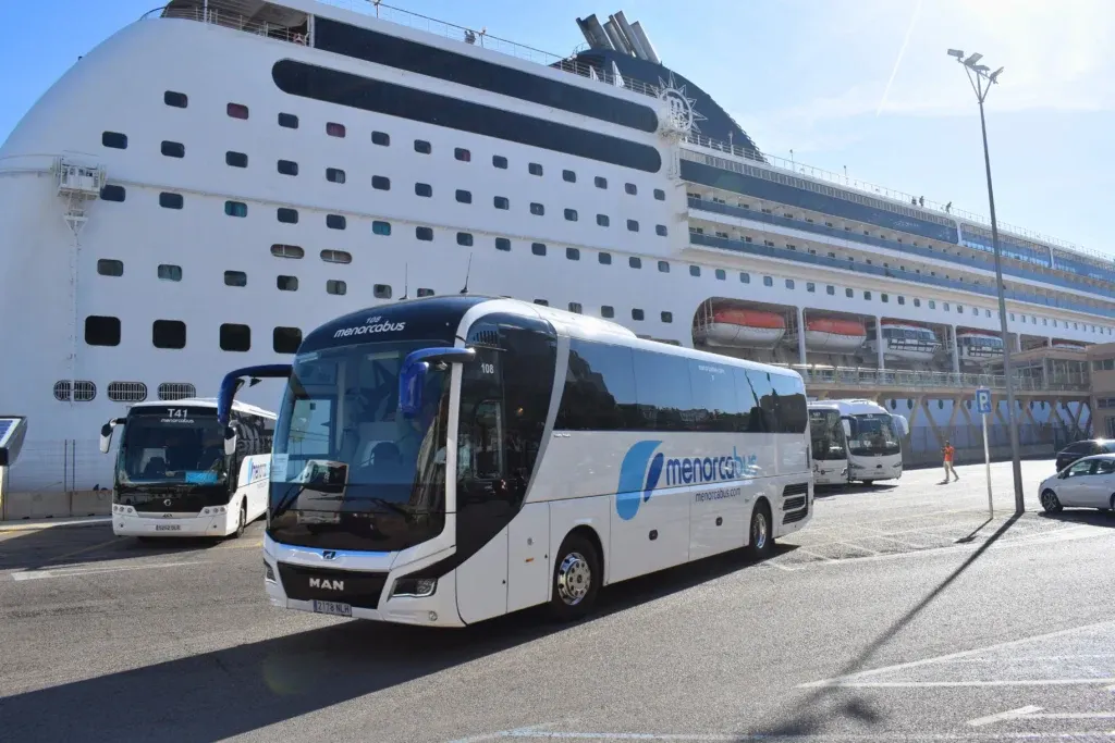 Autocar de Menorca Bus junto a un gran crucero atracado en el puerto de Maó.