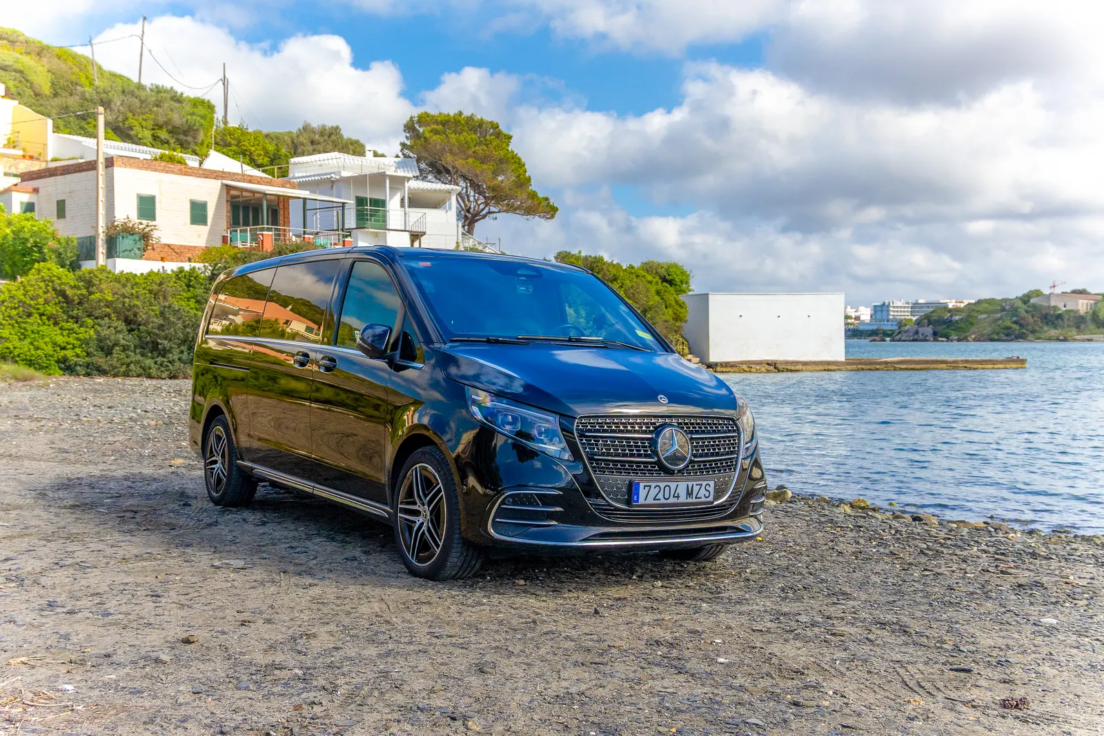 Mercedes Clase V negro de Menorca Bus aparcado junto a una cala con casas blancas al fondo.