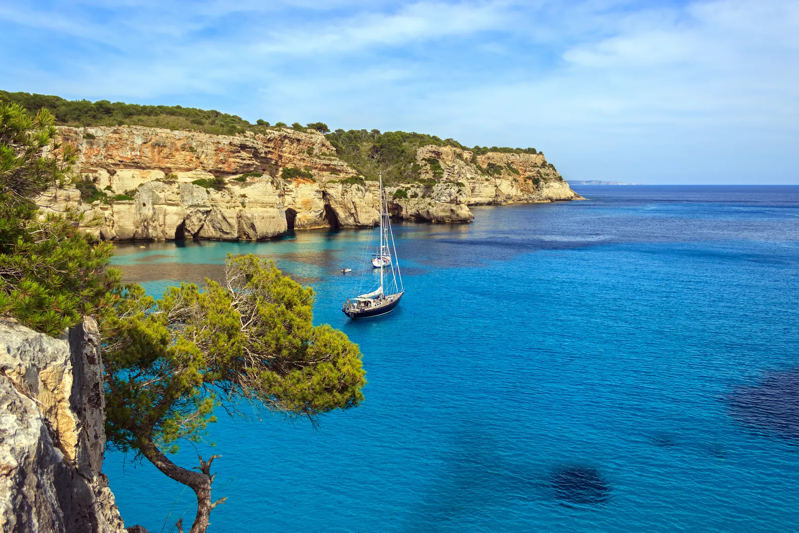 A boat at anchor on calm, turquoise water in the north of Menorca, seen from the sea.