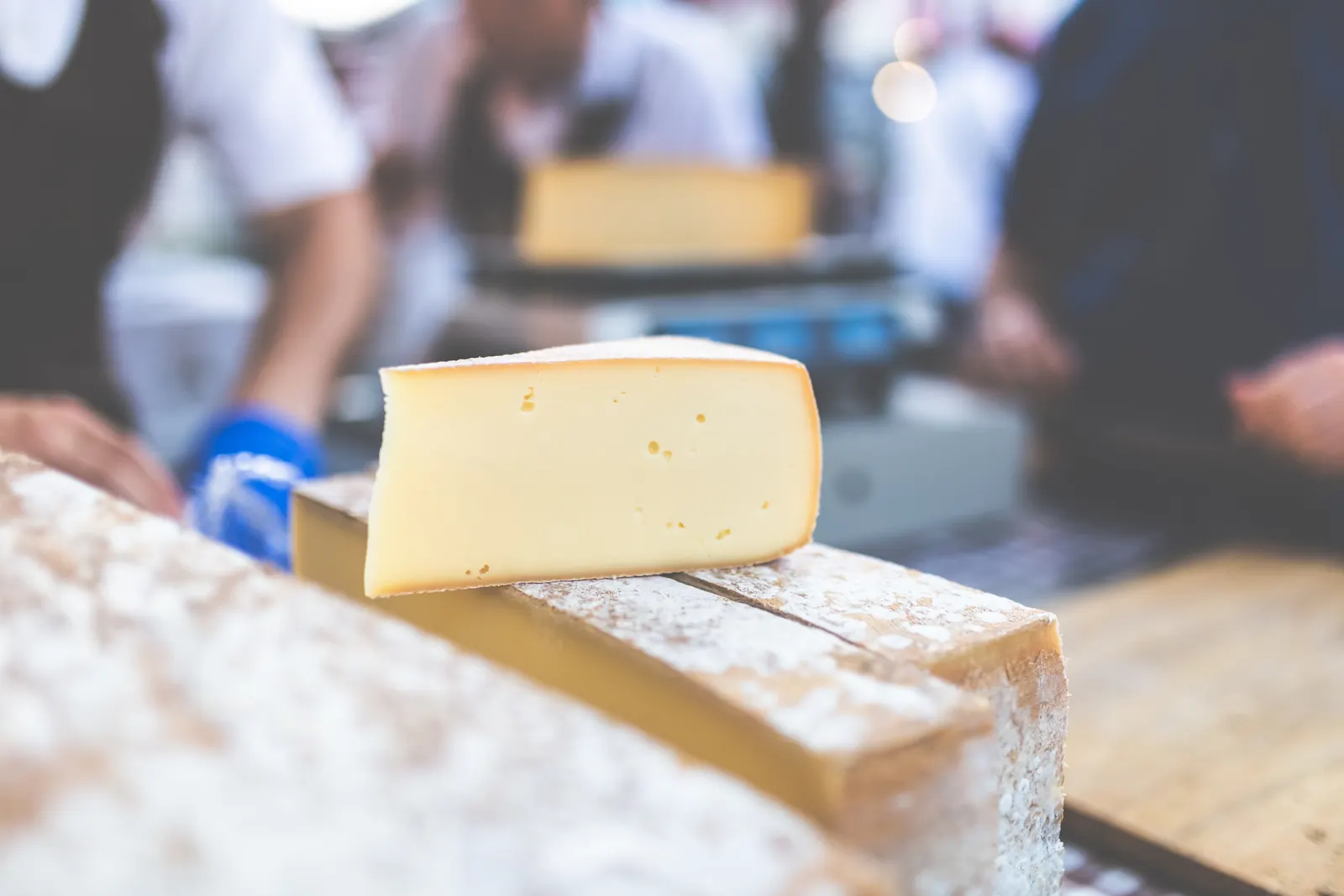 A wedge of aged cheese on a market stall counter, with local produce behind.