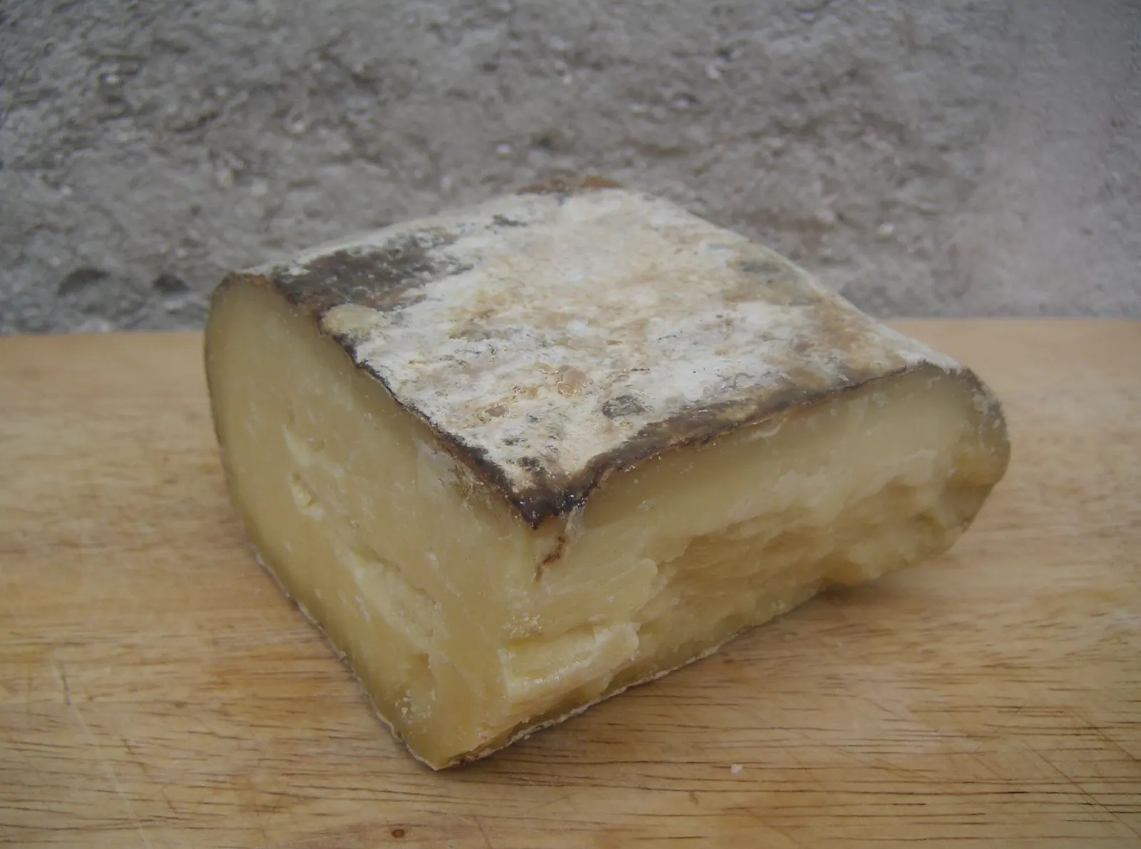 Bloque cuadrado de queso de Mahón-Menorca curado artesano sobre una tabla de madera; la cara superior conserva la corteza natural con moho blanco y grisáceo, y la pasta cortada se ve marfil y quebradiza.