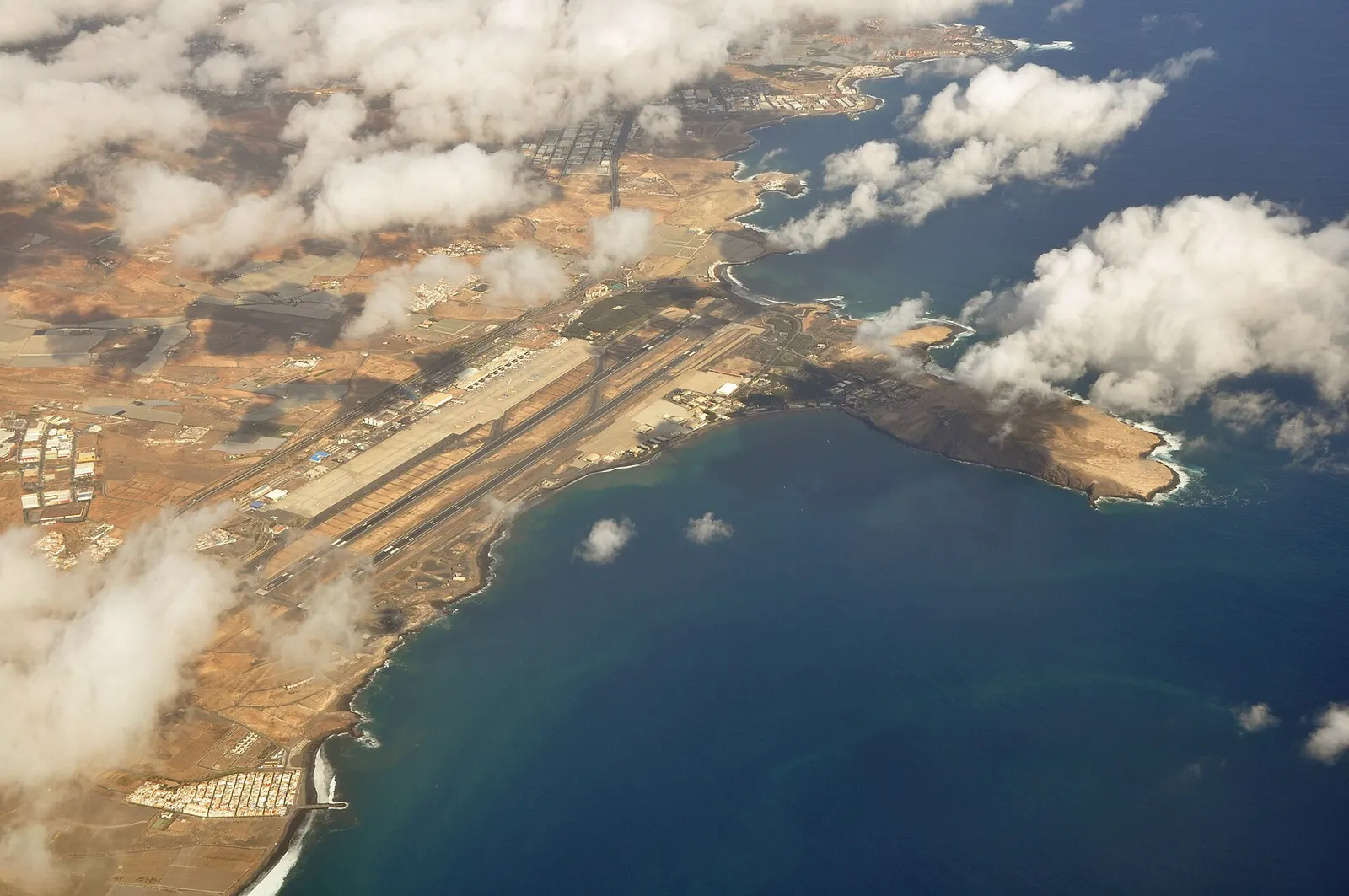 La costa este de Gran Canaria vista desde el aire, con la pista del aeropuerto junto al mar y nubes blancas sobre la bahía.