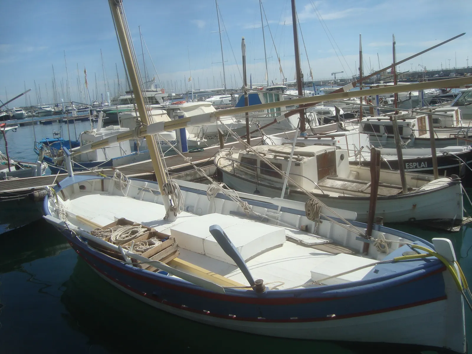 Un llaüt tradicional menorquín, de casco blanco y azul y vela latina recogida, amarrado en un puerto deportivo.