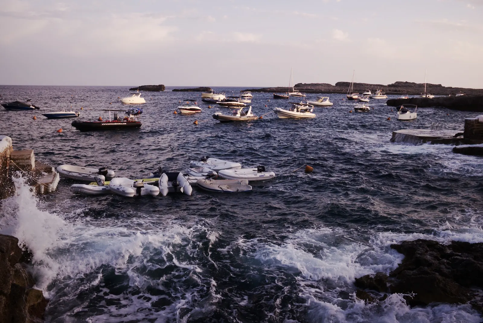 El mar rompiendo contra las rocas frente a Binibèquer, con embarcaciones fondeadas al fondo, al caer la tarde.