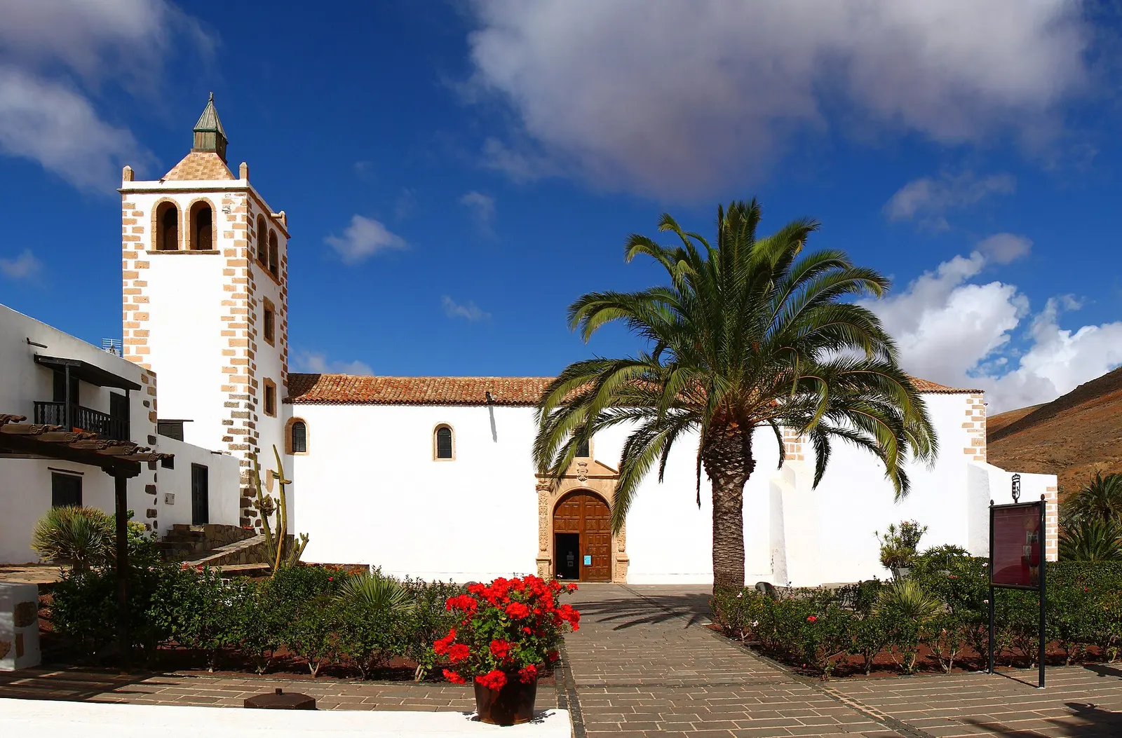 La iglesia de Santa María de Betancuria, blanca y con torre de cantería, tras una plaza con palmera y geranios rojos.
