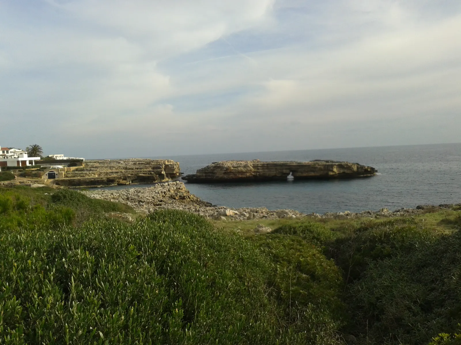 La costa rocosa de Cala Alcaufar, en Sant Lluís (Menorca), con casas blancas asomadas a un mar en calma.