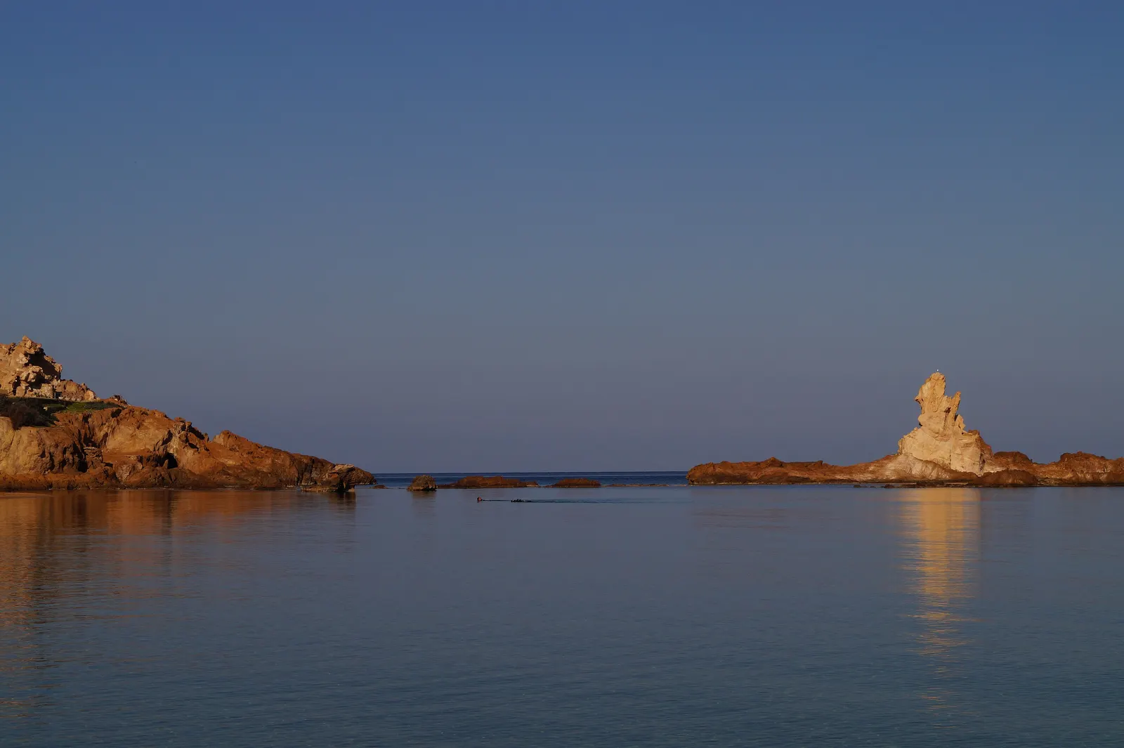 Cala Pregonda en hora dorada: el mar en calma refleja la luz cálida junto al islote de roca.