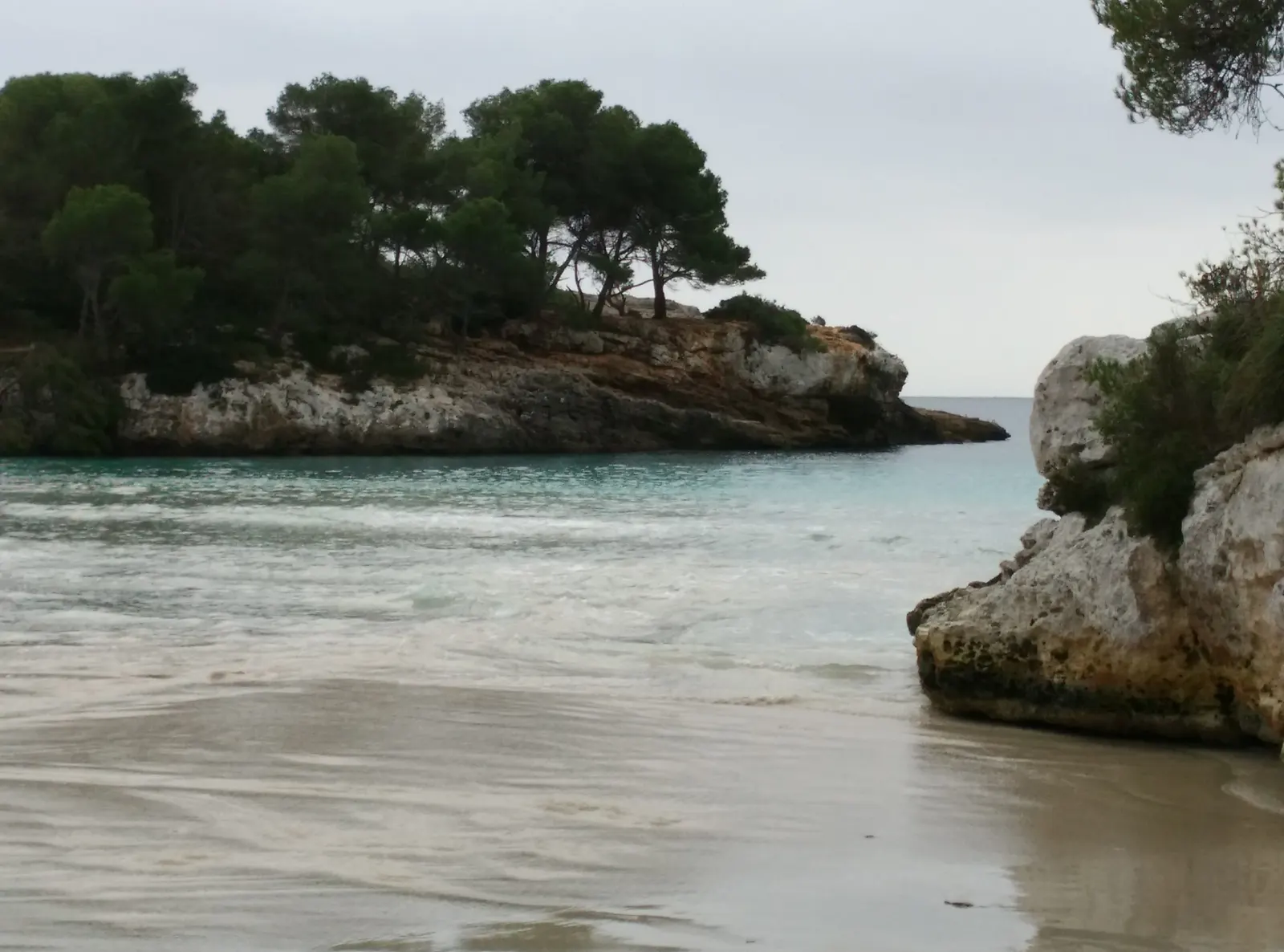 Cala en Turqueta con el cielo cubierto, la arena vacía y el promontorio de roca clara sobre el agua.