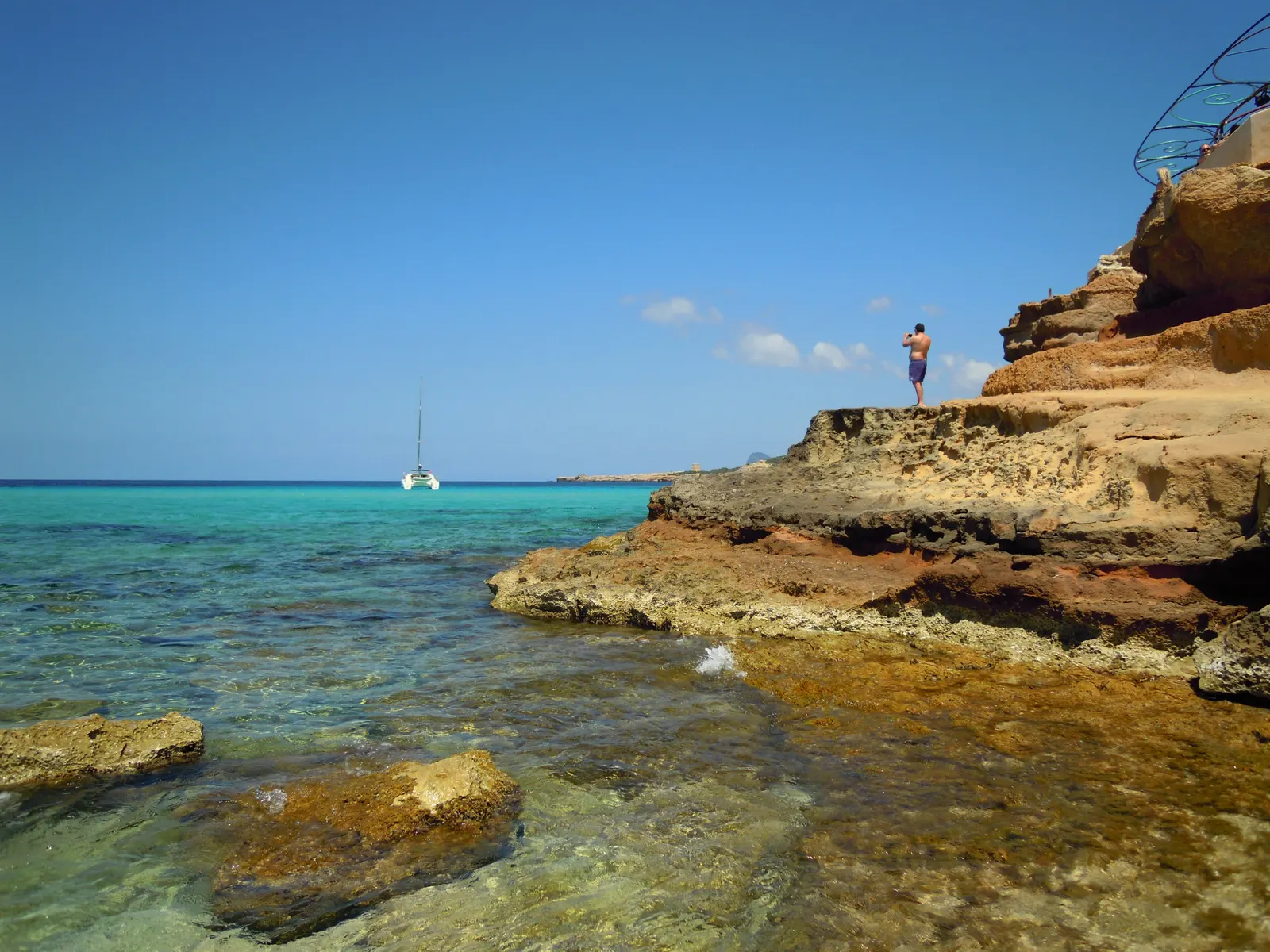 Cala Comte, en la costa de poniente de Ibiza: rocas doradas y agua turquesa, con un velero fondeado al fondo.