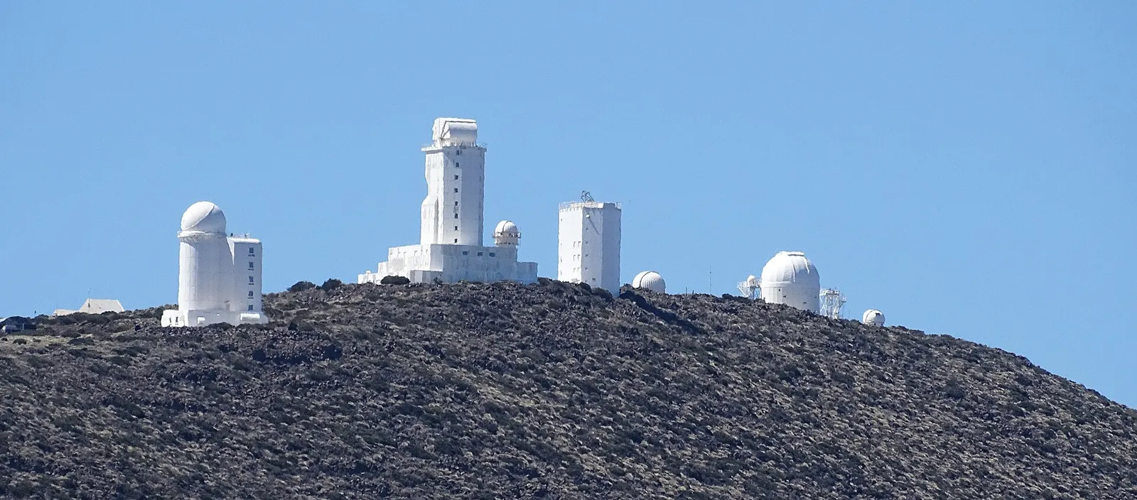 Las cúpulas y torres blancas del Observatorio del Teide sobre una loma volcánica oscura, bajo el cielo azul.