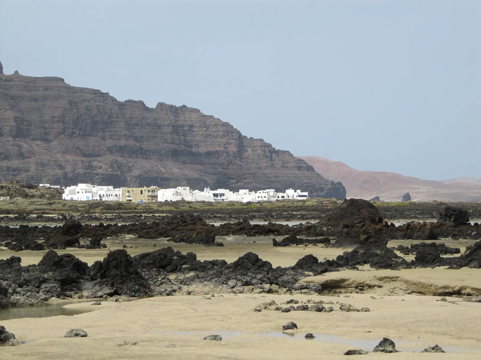 Die weißen Häuser von Órzola unter der Steilwand, von einem kleinen Strand zwischen Lavafelsen aus gesehen.