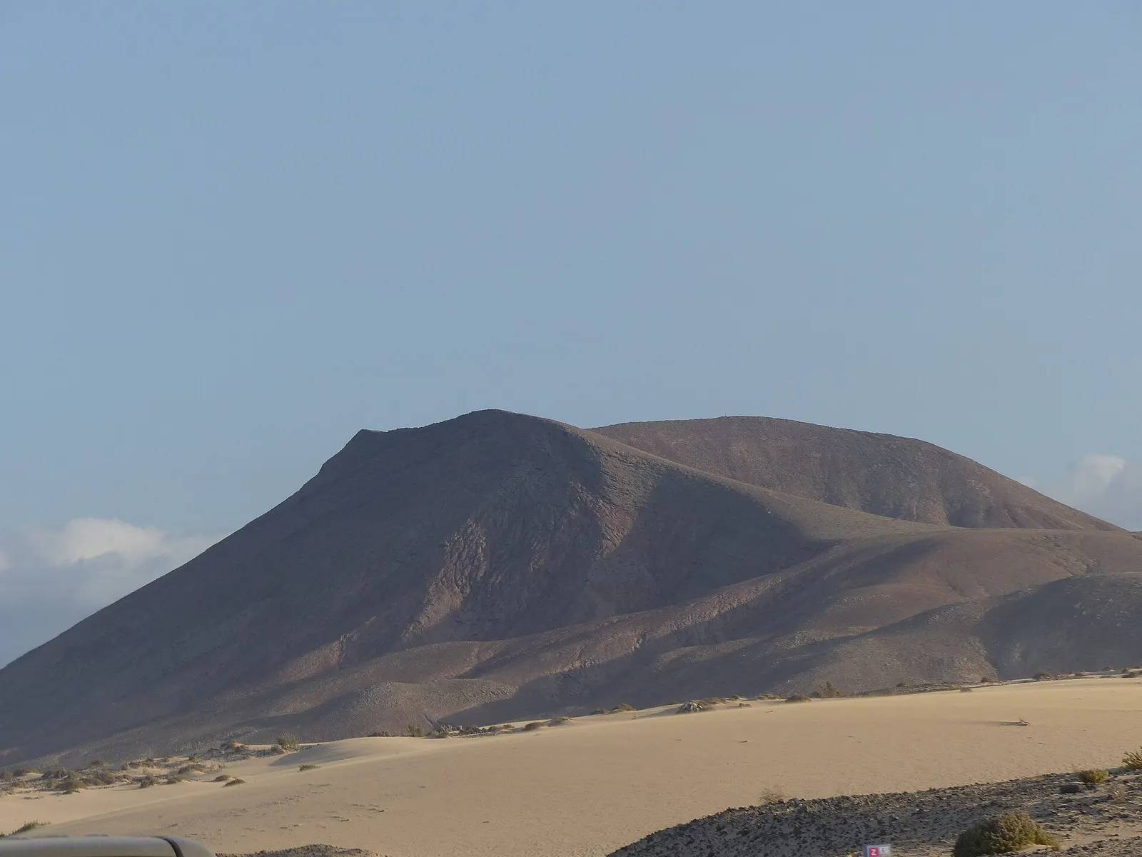 Una montaña volcánica de laderas rojizas se alza sobre la arena clara de las Dunas de Corralejo bajo un cielo sereno.