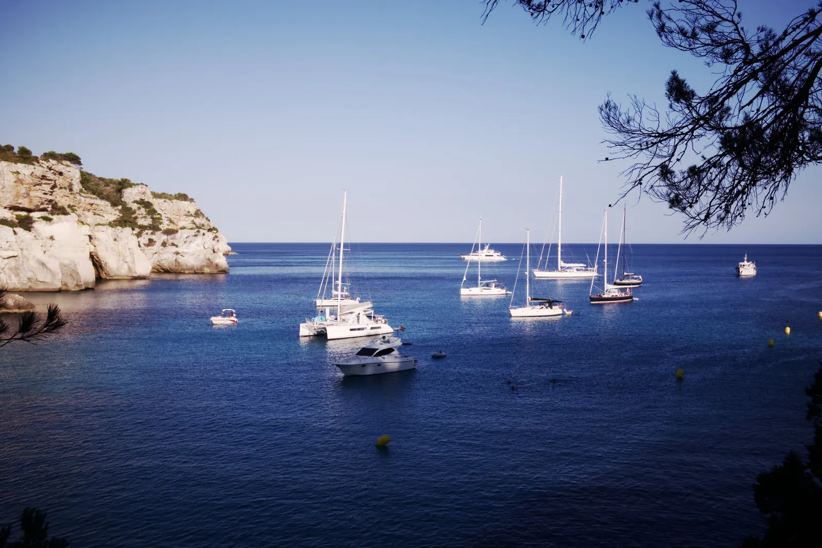 Varios veleros fondeados en una cala de aguas turquesas cerca de Cala Macarella, vista entre pinos desde un mirador de la costa sur.