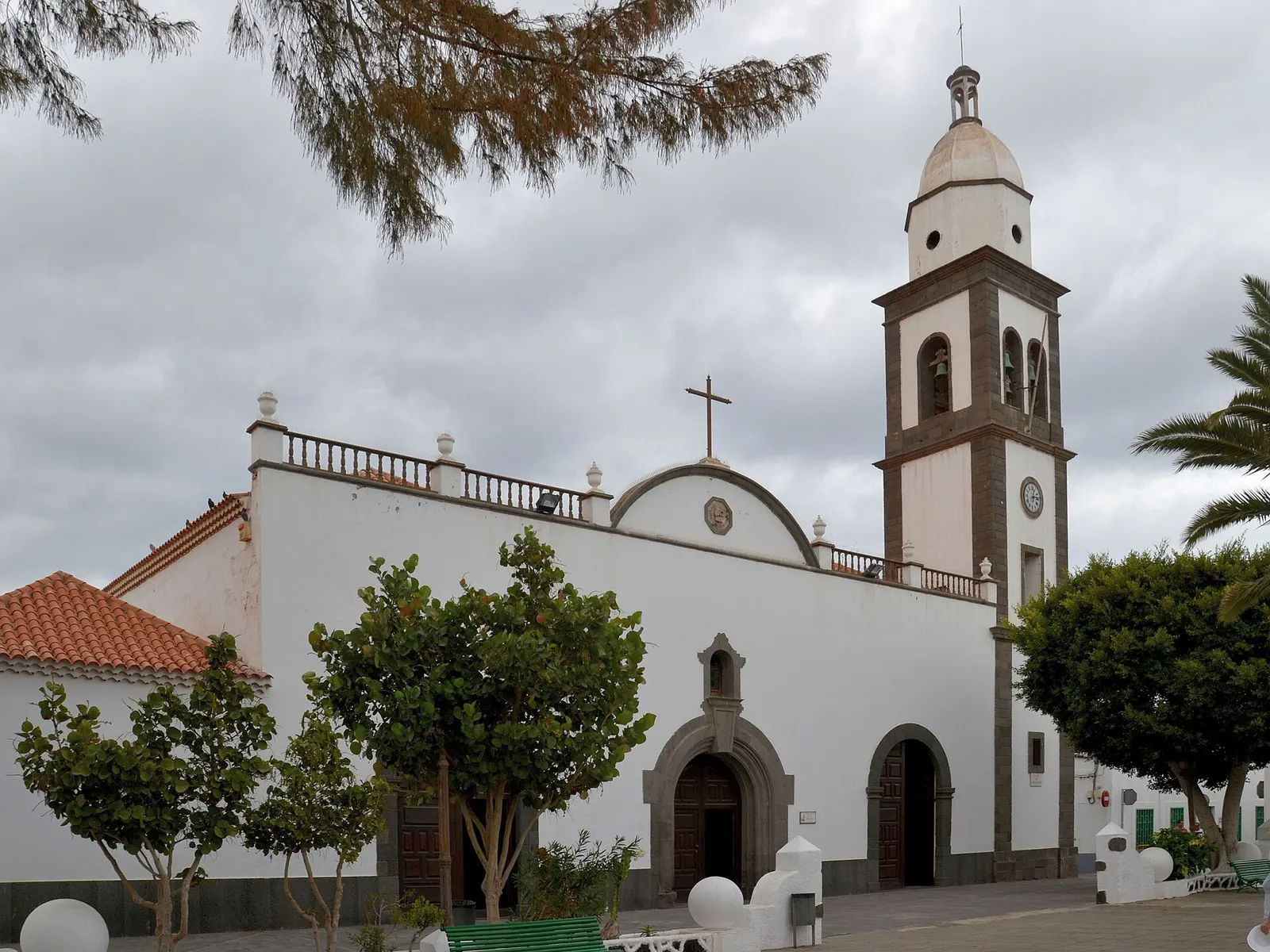 Die weiße Kirche San Ginés in Arrecife mit ihrem dunklen Steinturm zwischen Bäumen und Palmen.