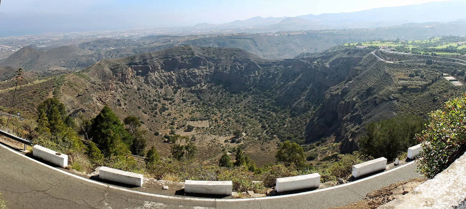 La caldera de Bandama desde su borde, un cráter verde y profundo, con las calles de un campo de golf asomando en lo alto a la derecha.