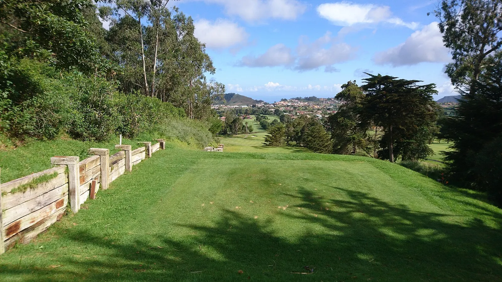 Una calle de golf verde entre pinos y eucaliptos, con un pueblo y montañas al fondo bajo el cielo azul.