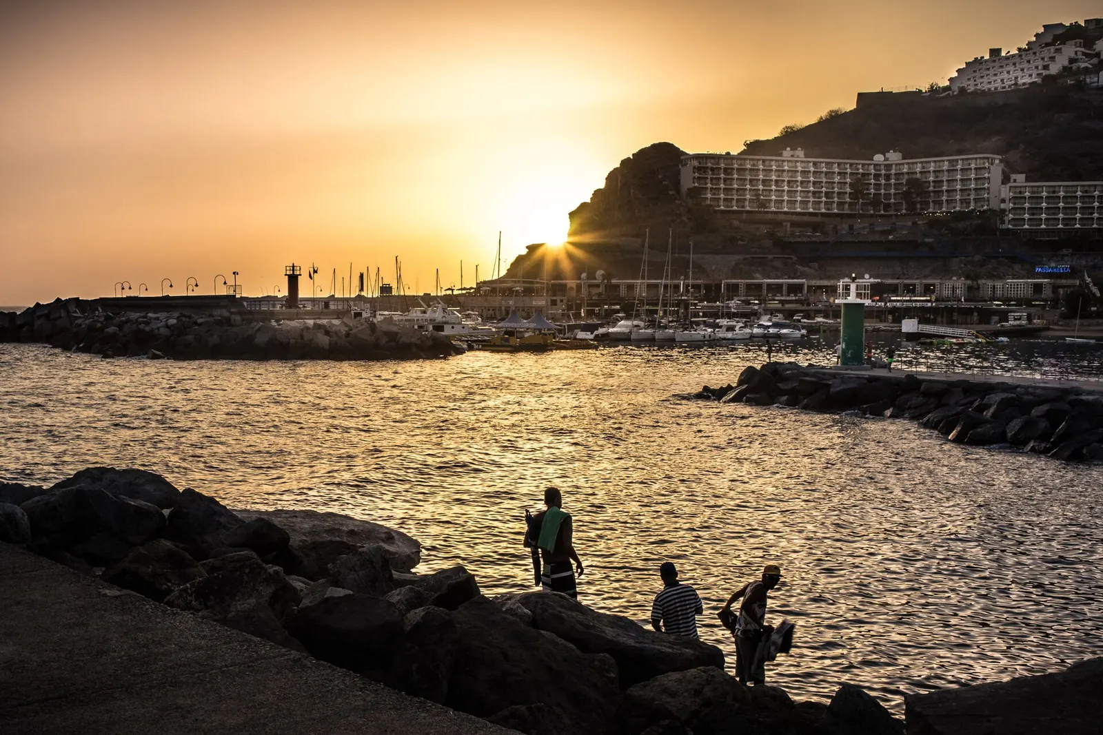 Atardecer dorado sobre la bocana de Puerto Rico, con el sol tras la ladera y tres siluetas mirando el mar desde las rocas.